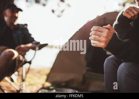 Nahaufnahme, Porträt einer Hand einer Frau mit Kaffee auf Campingplatz mit einem Mann im Hintergrund, beide sitzen vor dem Zelt. Stockfoto