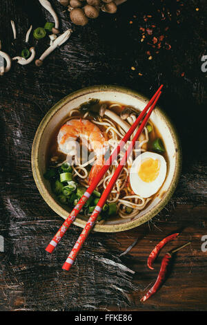 Keramikschale asiatischen Ramen-Suppe mit Garnelen, Nudeln, Zwiebeln, in Scheiben geschnitten, Ei und Pilzen, serviert mit roten Stäbchen und Chili pep Stockfoto