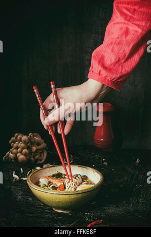 Weibliche Hand im roten Hemd nehmen durch rote Stäbchen aus asiatischen Ramen Nudeln Suppe mit Garnelen, Zwiebeln, in Scheiben geschnittenen Ei und Pilze, serv Stockfoto