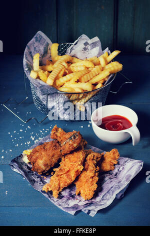 Chicken Pommes Streifen und Beine auf dem Papier mit Korb mit Pommes Frites und Schale mit Ketchup-Sauce über blaue Holztisch. Türkis Stockfoto