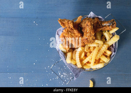 Pommes frites Hähnchenstreifen und Beine mit Pommes frites im Metallkorb über blaue Holztisch mit Meersalz. Ansicht von oben Stockfoto