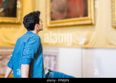 Eine halbe Stelle erschossen von ein nachdenklich hübscher junger Mann, Blick auf Malerei, sitzen auf Bank in einem Museum Stockfoto