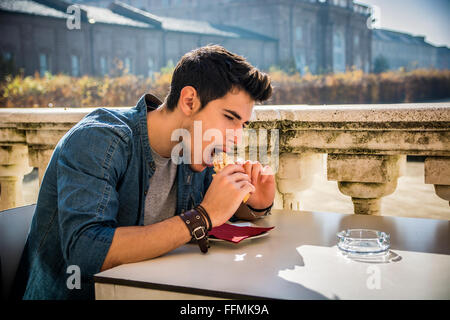 Junger Mann Betrieb Sandwich beim Sitzen im Café-Terrasse, Blick auf einer Seite ernst. Im Sommer Stockfoto