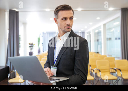 Porträt von Ernst attraktive Geschäftsmann stehen und hält Laptop im leeren Sitzungssaal Stockfoto