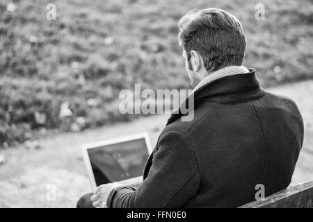 Schöne elegante Geschäftsmann sitzt auf einer Holzbank, arbeiten im Freien in einem städtischen Park, die Eingabe von Informationen auf seinem Laptop comp Stockfoto