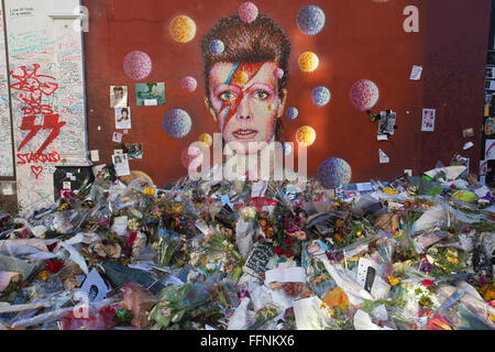 Blumen unter einem David Bowie "Aladdin Sane" Wandbild auf einer Straße Wand in Brixton, nach seinem Tod in einem behelfsmäßigen Schrein gemacht. Stockfoto
