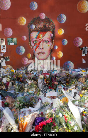 Blumen unter einem David Bowie "Aladdin Sane" Wandbild auf einer Straße Wand in Brixton, nach seinem Tod in einem behelfsmäßigen Schrein gemacht. Stockfoto