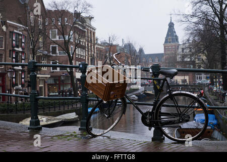 Fahrrad angekettet an Geländer an einem Kanal in Amsterdam, die Niederlande, auf einem nassen und regnerischen Tag im winter Stockfoto