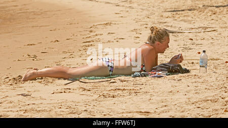 Junge blonde Frau im Bikini auf Sand am Strand mit Mobiltelefon mit Flasche Wasser in der Nähe in Australien Stockfoto