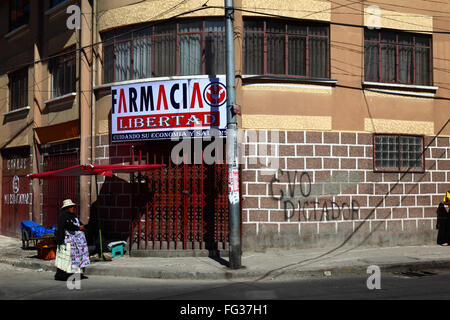 Aymara Frau, die an Evo Diktator / Evo Diktator Graffiti an der Wand von Farmacia Libertad / Liberty or Freedom Pharmacy Gebäude, La Paz, Bolivien vorbeigeht Stockfoto