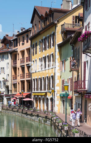 Menschen zu Fuß vorbei an Häusern und eine Frau ruht auf einem Balkon in der Altstadt von Annecy am Fluss Thiou an einem sonnigen Morgen Stockfoto