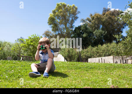 Kind Junge mit Kamera im freien dabei sein erste Foto in der Wiese sitzen und genießen sonnigen Tag in der Natur. Stockfoto