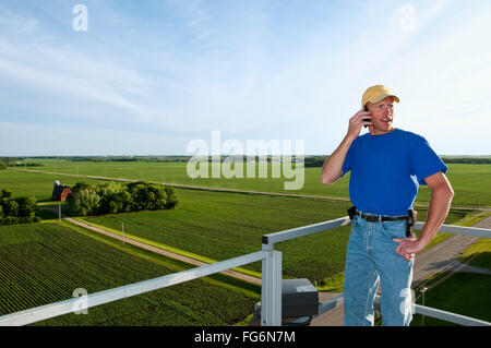 Landwirtschaft - spricht ein junger Landwirt auf seinem Handy auf ein Silo mit einem Hof und Felder im Hintergrund / in der Nähe von Northrop, Minnesota, USA. Stockfoto