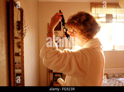 Applying Reife Frau macht ihr Haar morgens mit einer beheizten Curling Zange im Schlafzimmer Stockfoto