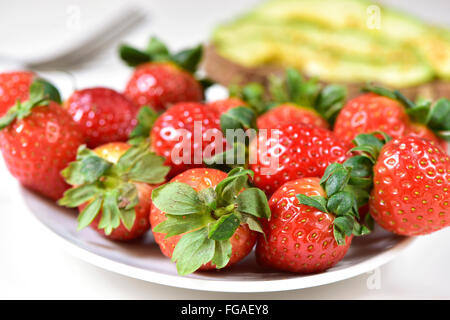 Nahaufnahme von einem weißen Keramik Teller voll mit leckeren Erdbeeren und eine Avocado-Toast im Hintergrund auf einem gedeckten Tisch Stockfoto
