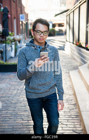 Junge asiatische Mann trägt Brille und hält Smartphone zu Fuß auf Stadtstraße Stockfoto