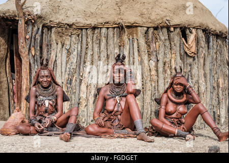 Drei Himba-Frauen sitzen auf dem Boden vor einer Holzhütte. Stockfoto