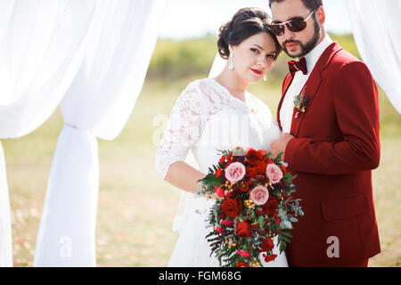 Extravagante Braut und Bräutigam, schönes paar Hochzeit Foto zu schießen. Mann im roten Anzug, Sonnenbrille mit Fliege. Sommer sonniger Tag im Freien, Arch. Stockfoto