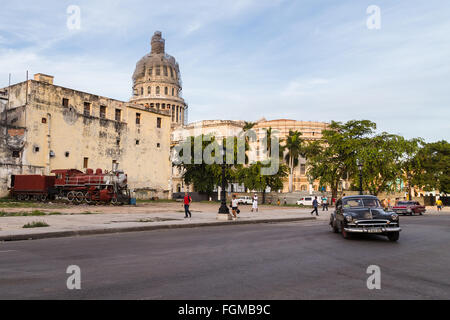 Eine Reihe von Bäumen, eine Straße von Altautos & eine alte Dampflok vor dem Kapitol in Havanna gesehen. Stockfoto