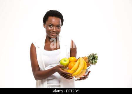 Schöne fröhliche afrikanische amerikanische junge Frau stehen und halten Glasschale mit exotischen Früchten auf weißem Hintergrund Stockfoto