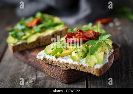Gesunden Toast mit Avocado, Rucola, Ziegenkäse und Sonne getrockneten Tomaten auf rustikalen Holztisch. Selektiven Fokus Stockfoto