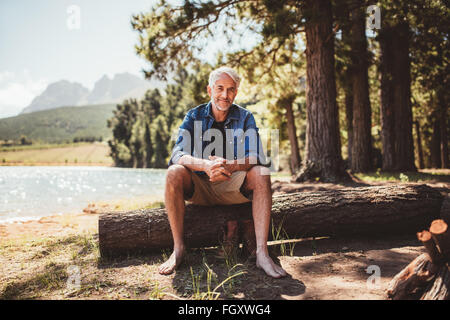 Porträt des entspannten reifer Mann sitzt auf einem Holzscheit in der Nähe des Sees. Reifer Mann genießt einen Tag am See. Stockfoto
