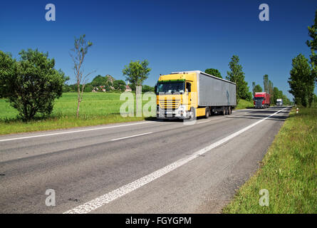 Straße, gesäumt von Bäumen in einer ländlichen Landschaft, farbige drei vorbeifahrenden LKW. Grüne Felder, Dörfer im Hintergrund. Stockfoto