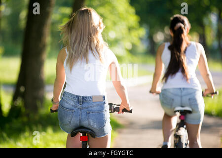 Zwei junge schöne Frauen Freundinnen tragen Jeans Shorts Radfahren auf Bürgersteig auf der Straße an sonnigen Sommertag, Rückansicht Stockfoto