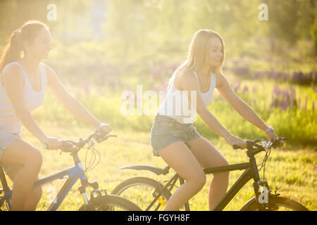 Zwei süße junge glücklich lächelnd schöne Frauen Freundinnen tragen Jeans Shorts Fahrradfahren im Park in hellem Sonnenlicht im Sommer Stockfoto