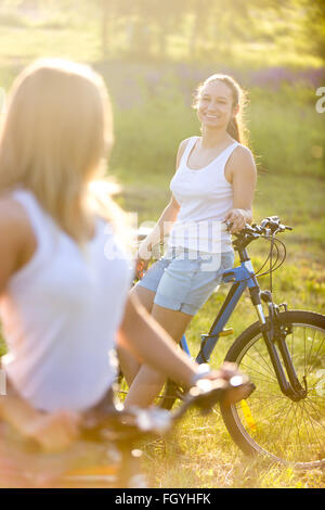 Zwei fröhliche schönen Teenager Freundinnen tragen lässige Jeans Hosen sitzen auf dem Fahrrad im Park an sonnigen Sommertag Stockfoto