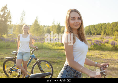 Zwei schöne Radfahrer Freundinnen tragen lässige weiße Tank-Tops und Jeans-Shorts mit Fahrrädern im Natur Park stehen Stockfoto