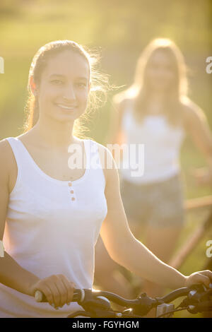 Zwei schöne Mädchen im Teenageralter mit Fahrrädern tragen lässige weiße Tank-Tops und Jeans-Shorts im Natur Park auf hell stehen Stockfoto