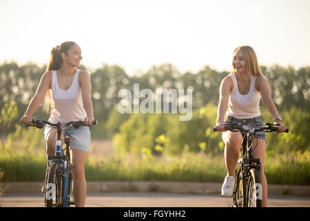 Zwei schöne junge Frauen Reiten Fahrräder tragen lässige weiße Tank-Tops und Jeans-Shorts im Park auf hellen sonnigen Sommertag Stockfoto