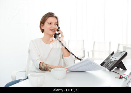 Lächelnde schöne junge Frau sitzt im Büro und am Telefon sprechen Stockfoto