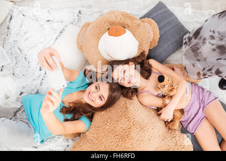 Draufsicht der zwei glücklich schönen Schwestern auf großen Plüschbär und nehmen Selfie liegend mit Handy zu Hause Stockfoto