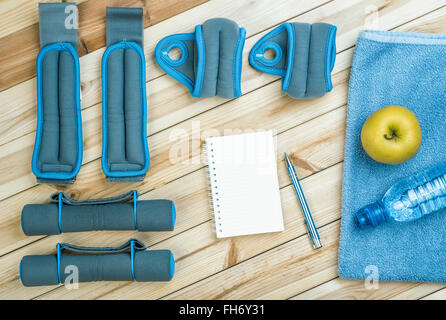 Sportgeräte. Hanteln, Fussblei, Handgelenk Gewichte, Handtuch, Apple, Flasche Wasser und Notebook zu Trainingsplan auf Brettern. Stockfoto