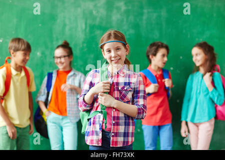 Hübsches Mädchen mit Blick in die Kamera mit ihren Mitschülern im Chat auf Hintergrund rucksack Stockfoto