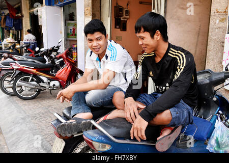 zwei Personen, die auf einem geparkten Roller entlang einer belebten städtischen Straße sitzen, die mit anderen Motorrädern gesäumt ist - Straßen von Ho-Chi-Minh-Stadt, Vietnam Stockfoto