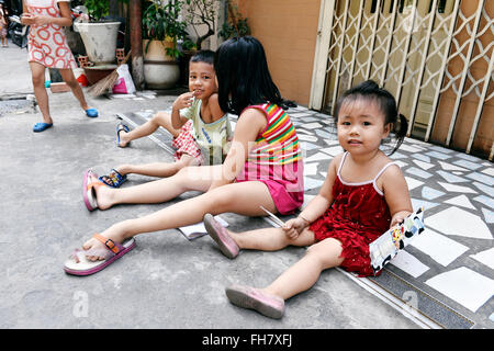 Kinder, die auf einem gepflasterten Außengelände in der Nähe eines Gebäudeeingangs sitzen und spielen - Straßen von Ho-Chi-Minh-Stadt, Vietnam Stockfoto