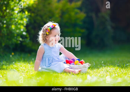 Lustige kleine Mädchen spielen im sonnigen Garten. Baby auf Ostereiersuche. Kleinkind Kind mit weißen Korb Kommissionierung bunte Eiern Stockfoto