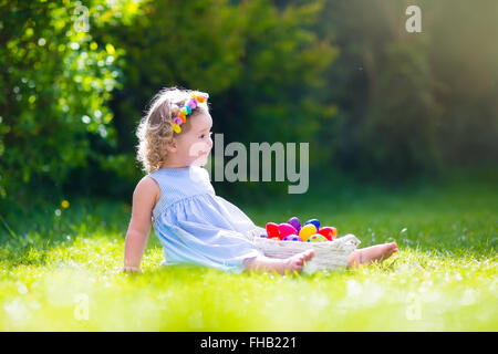 Lustige kleine Mädchen spielen im sonnigen Garten. Baby auf Ostereiersuche. Kleinkind Kind mit weißen Korb Kommissionierung bunte Eiern Stockfoto