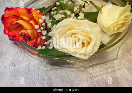 Drei schöne Rosen liegen auf einer Glasschale auf dem Tisch, bedeckt mit einer weißen Tischdecke. Stockfoto