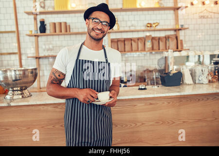 Porträt von glücklichen jungen Barista bei der Arbeit. Kaukasischen Mann mit Schürze und Mütze stand vor Café Zähler mit Tasse Kaffee Stockfoto