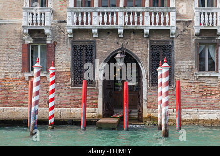 Anlegestelle eines alten Backsteingebäudes mit einem gewölbten Eingang auf dem Canal Grande, Venedig, mit rot und rot und weiß gestreiften Anlegestellen Stockfoto