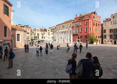 Sonnendurchflutete Gebäude in warmen Farben und ein Markt im Freien auf dem Campo Sant'Angelo, Venedig, in dem sich Gruppen von Touristen unter blauem Himmel tummeln Stockfoto