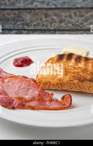 Zwei Speckscheiben Speck und eine Scheibe Toast auf einem Teller mit Butter und Tomaten Ketchup auf einem Tisch Stockfoto