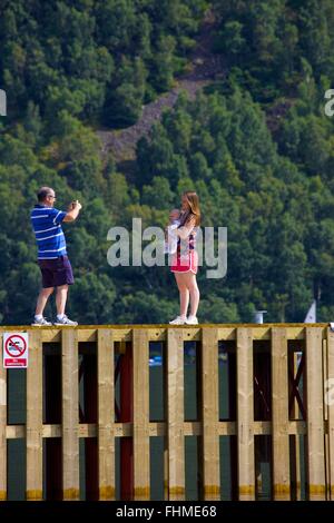 Lake District National Park. Mann nehmen Foto von seiner Frau und Baby mit Ihrem Smartphone auf einem Steg in Lake Ullswater. Stockfoto