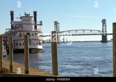 Malerische Schuss von einem mehrstufigen Ausflugsboot in der Nähe von der Wasser-Straße-Promenade am Cape Fear River mit einer Brücke in der Ferne Stockfoto