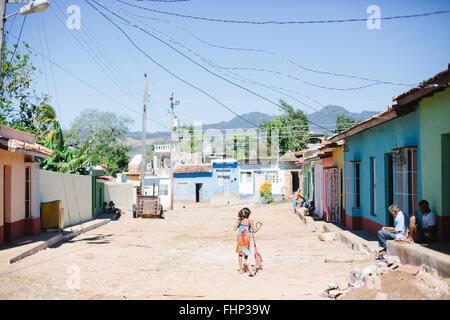 In einer armen Gegend von Trinidad in Kuba schiebt eine Mädchen ihrem Roller auf der unbefestigten Straße, gesäumt von bunten Häusern Stockfoto
