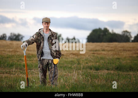 Mann mit Metalldetektor und Schaufel posiert im Feld. Schatzsucher trägt Tarnung und sieht aus wie zuversichtlich Stockfoto